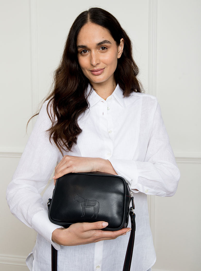 Woman wearing a large premium bridle butt navy leather Bridget crossbody camera hand bag that has been hand stitched in England with red threading and embossed with the Newmarket 875 brand horse logo.