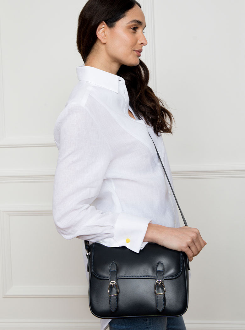 Woman wearing a large premium bridle butt navy leather crossbody Ceres hand bag that has been hand stitched in England with 2 front facing buckle straps and embossed with the Newmarket 875 horse logo on the back.
