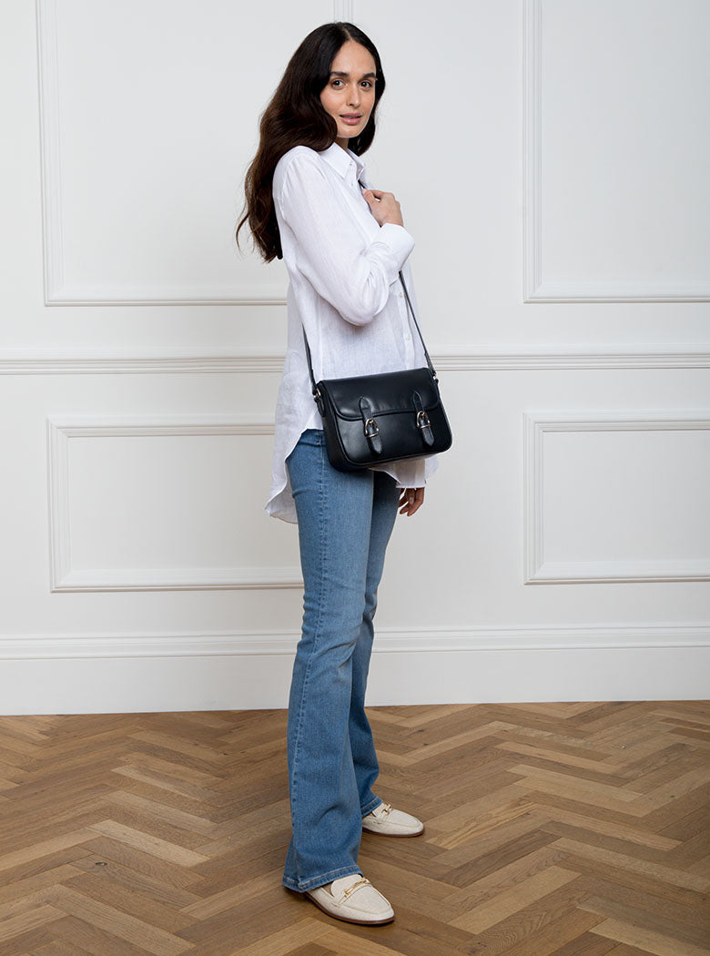 Woman wearing a large premium bridle butt navy leather crossbody Ceres hand bag that has been hand stitched in England with 2 front facing buckle straps and embossed with the Newmarket 875 horse logo on the back.