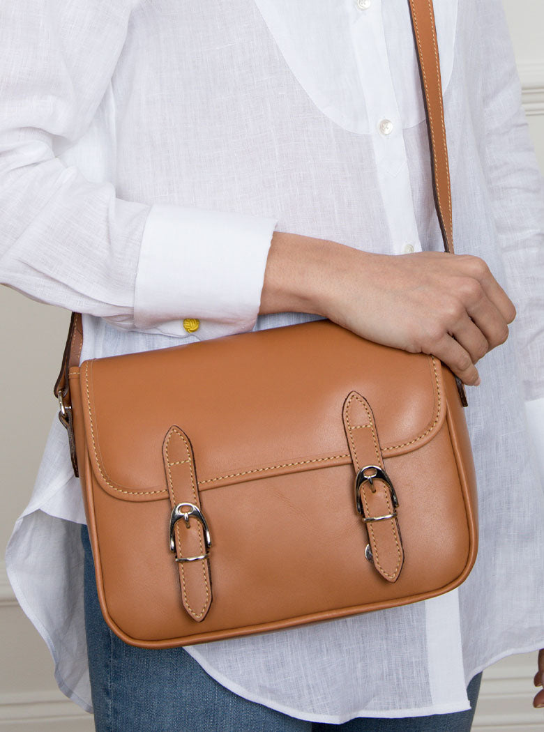Woman wearing a large premium bridle butt tan leather crossbody Ceres hand bag that has been hand stitched in England with 2 front facing buckle straps and embossed with the Newmarket 875 horse logo on the back.