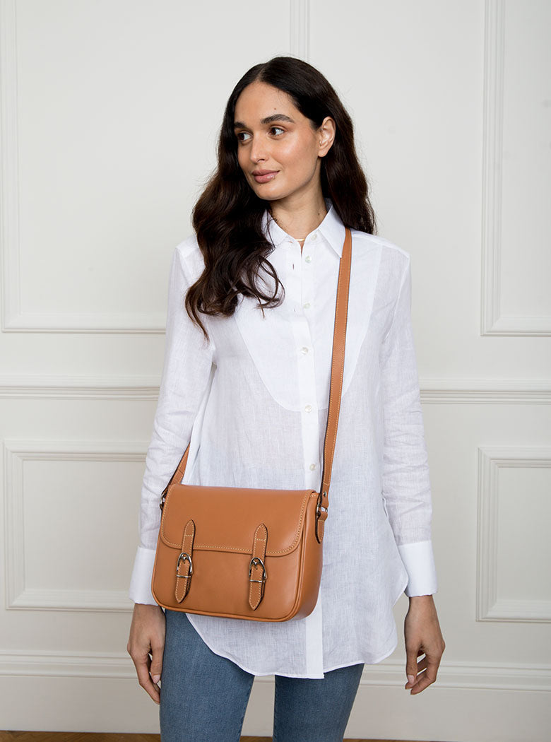 Woman wearing a large premium bridle butt tan leather crossbody Ceres hand bag that has been hand stitched in England with 2 front facing buckle straps and embossed with the Newmarket 875 horse logo on the back.