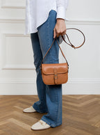 Woman wearing a small premium bridle butt tan leather crossbody Faith hand bag that has been hand stitched in England with 1 front facing buckle strap and the back embossed with Newmarket 875 horse logo.