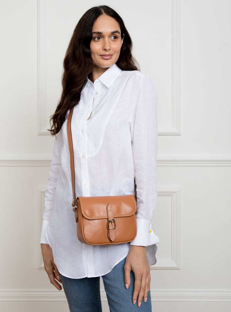Woman wearing a small premium bridle butt tan leather crossbody Faith hand bag that has been hand stitched in England with 1 front facing buckle strap and the back embossed with Newmarket 875 horse logo.
