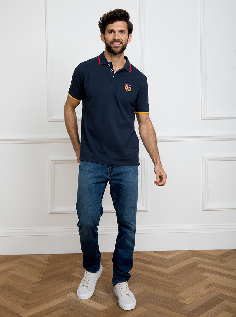 Man wearing navy polo shirt in 100% cotton with Newmarket 875 crest embroidered on the front and giant gold number 5 on the back with gold sleeve trimming and red collar trimming.