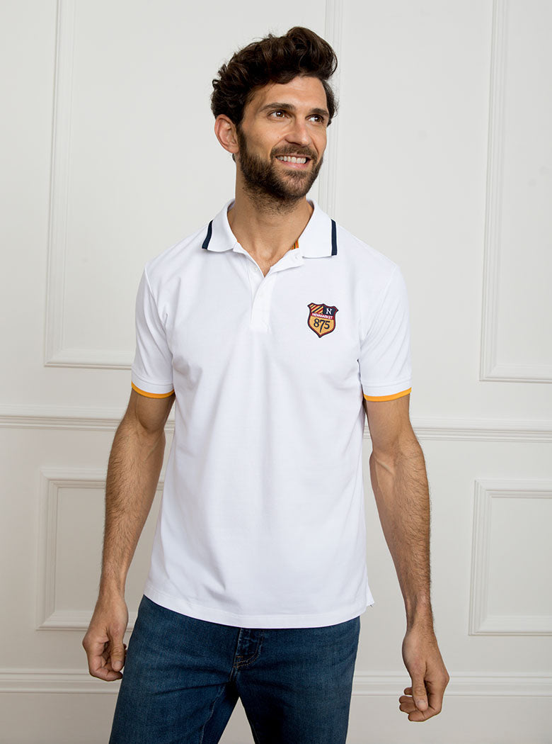 Man wearing white polo shirt in 100% cotton with Newmarket 875 crest embroidered on the front with navy sleeve trimming and gold collar trimming.
