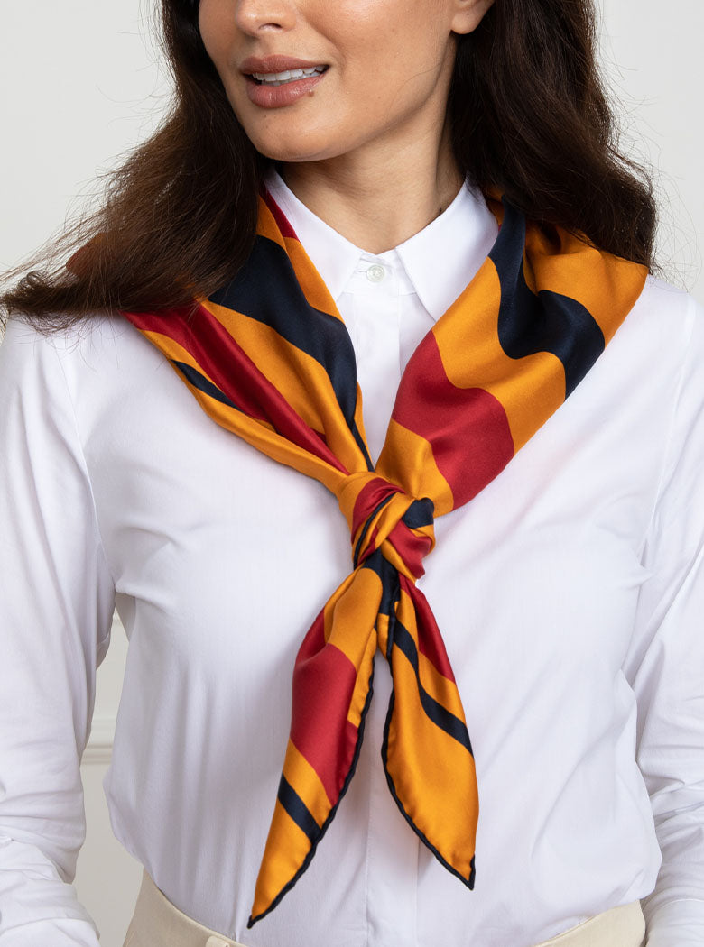Woman wearing 100% silk 90x90 foulard scarf in traditional Newmarket red, navy, gold yellow stripe with navy hand stitched border. 
